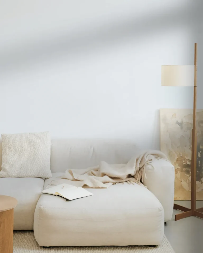 اتاق نشیمن مینیمال با مبل روشن، پتو و کوسن‌های بافتنی سفید و چراغ ایستاده چوبی